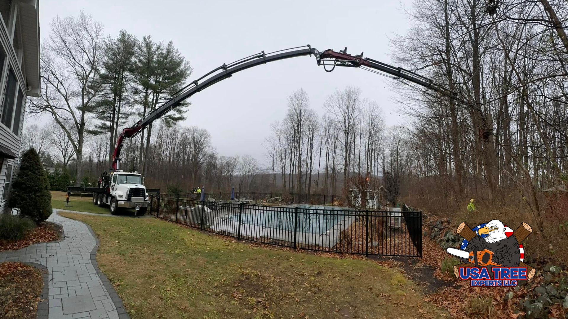 A large crane truck from a tree service company extends its hydraulic arm over a residential pool area to remove tree limbs from a wooded area in the backyard. A worker in high-visibility gear supervises the operation, demonstrating safe and precise tree removal near a home.